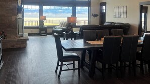 Un comedor espacioso con una mesa grande y sillas, una chimenea de piedra y amplios ventanales que ofrecen vistas a un lago y montañas.