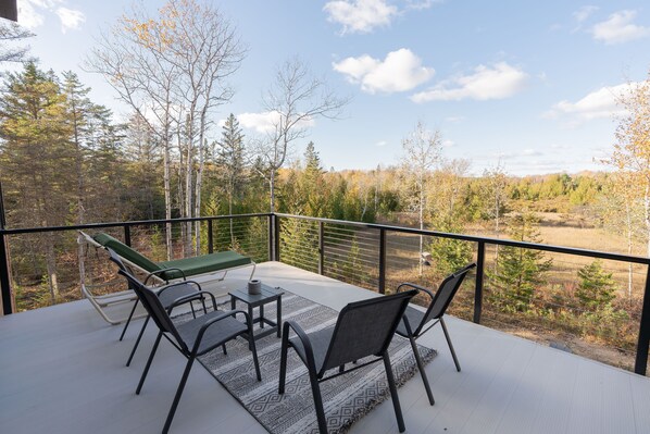 A balcony with a table, chairs, and a green lounge chair, overlooking a forested area.