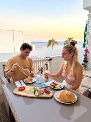 Una coppia cena all'aperto con vista sul mare, un tavolo imbandito di cibo e bevande, e una sedia in vimini.