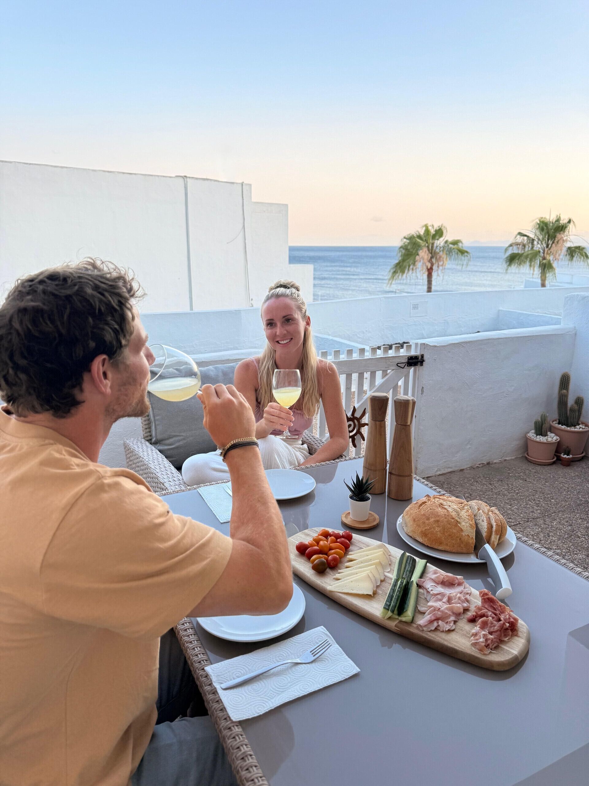 Ein Paar genießt ein Essen an einem Tisch im Freien mit Blick auf das Meer, mit verschiedenen Käsesorten und einer Weinflasche.