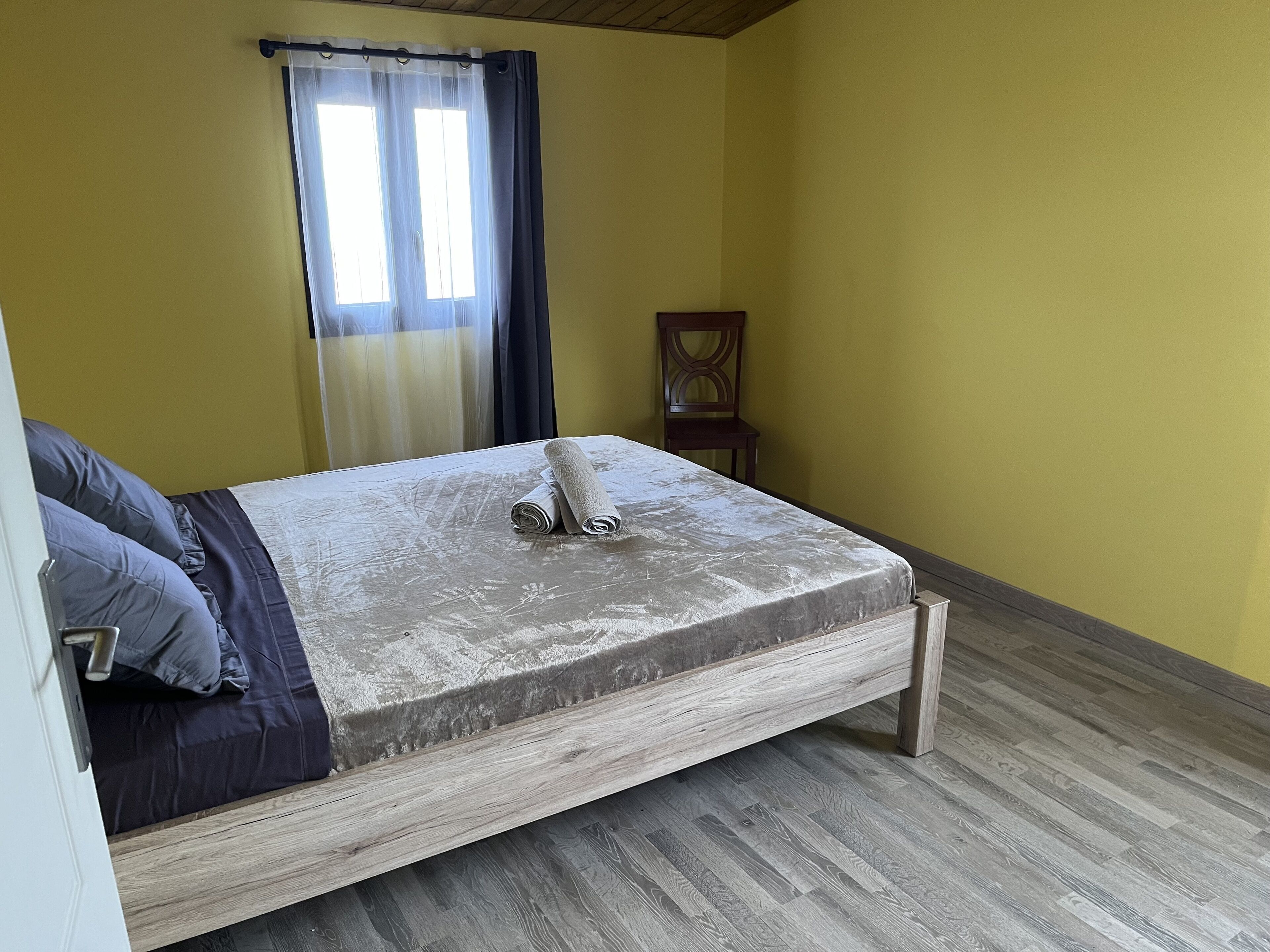 Ein Einzelbett mit Holzbettrahmen, einer Matratze und aufgerollten Handtüchern. Ein Stuhl und ein Fenster mit Vorhängen sind ebenfalls vorhanden.