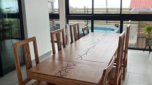Een eetgedeelte met een lange houten tafel en stoelen, grote ramen met uitzicht op een zwembad en een gebouw met een rood dak.
