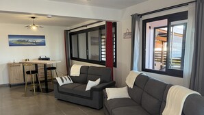 Een moderne woonkamer met een grijze bank, een eethoek met een houten tafel en barstoelen, en een groot raam met gordijnen.