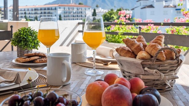 Ein Tisch mit Croissants, einer Obstschale und zwei Gläsern Orangensaft, mit Blick auf die Berge im Hintergrund.