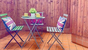 Een terras met een houten omheining, een ronde tafel, twee stoelen met Union Jack kussens en een bloempot.