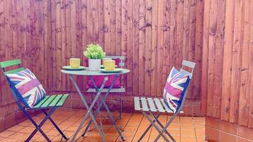 A patio with a wooden fence, a round table, two chairs with Union Jack cushions, and a potted plant.