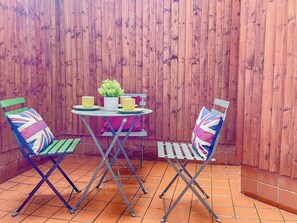 A patio with a wooden fence, a round table, two chairs with Union Jack cushions, and a potted plant. - 3-bed Pimlico Flat, Garden | victoria tate britain (Greater London)