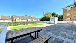 Un patio al aire libre con un banco de madera y suelo empedrado, rodeado de setos y edificios al fondo.