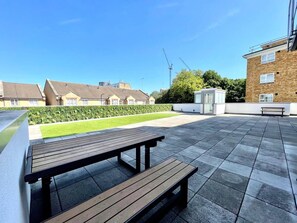 An outdoor patio area with a wooden bench and a paved floor, surrounded by a hedge and buildings in the background. - 3bdr Hoxton Flat Near Old st, shoreditch + balcony (London)