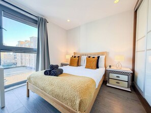A hotel room with a large bed, a nightstand, a lamp, and a view of the city through a window.