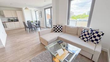 A modern living room with a glass coffee table, a sofa with checkered cushions, and a view of the city through large windows.