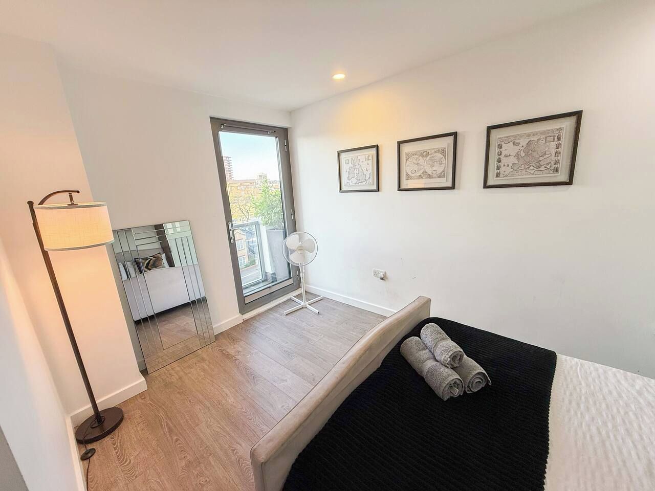 Un dormitorio con cama, mesita de noche, espejo, lámpara de pie y vistas al exterior.