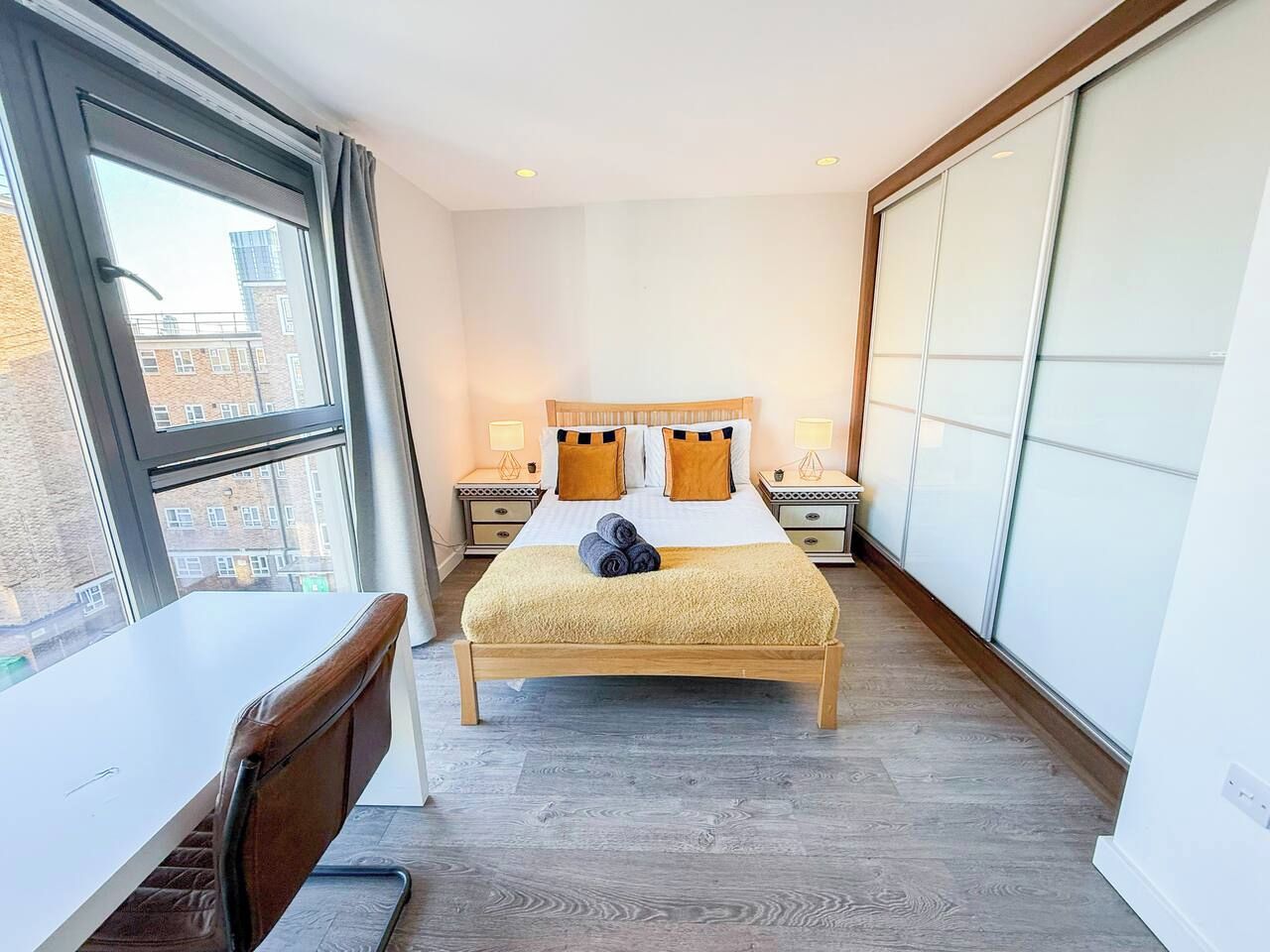 A hotel room with a bed, bedside tables, a desk, and a large window.