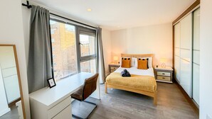 Quarto de hotel com uma cama, escrivaninha, cadeira, mesa de cabeceira e uma janela com cortinas.