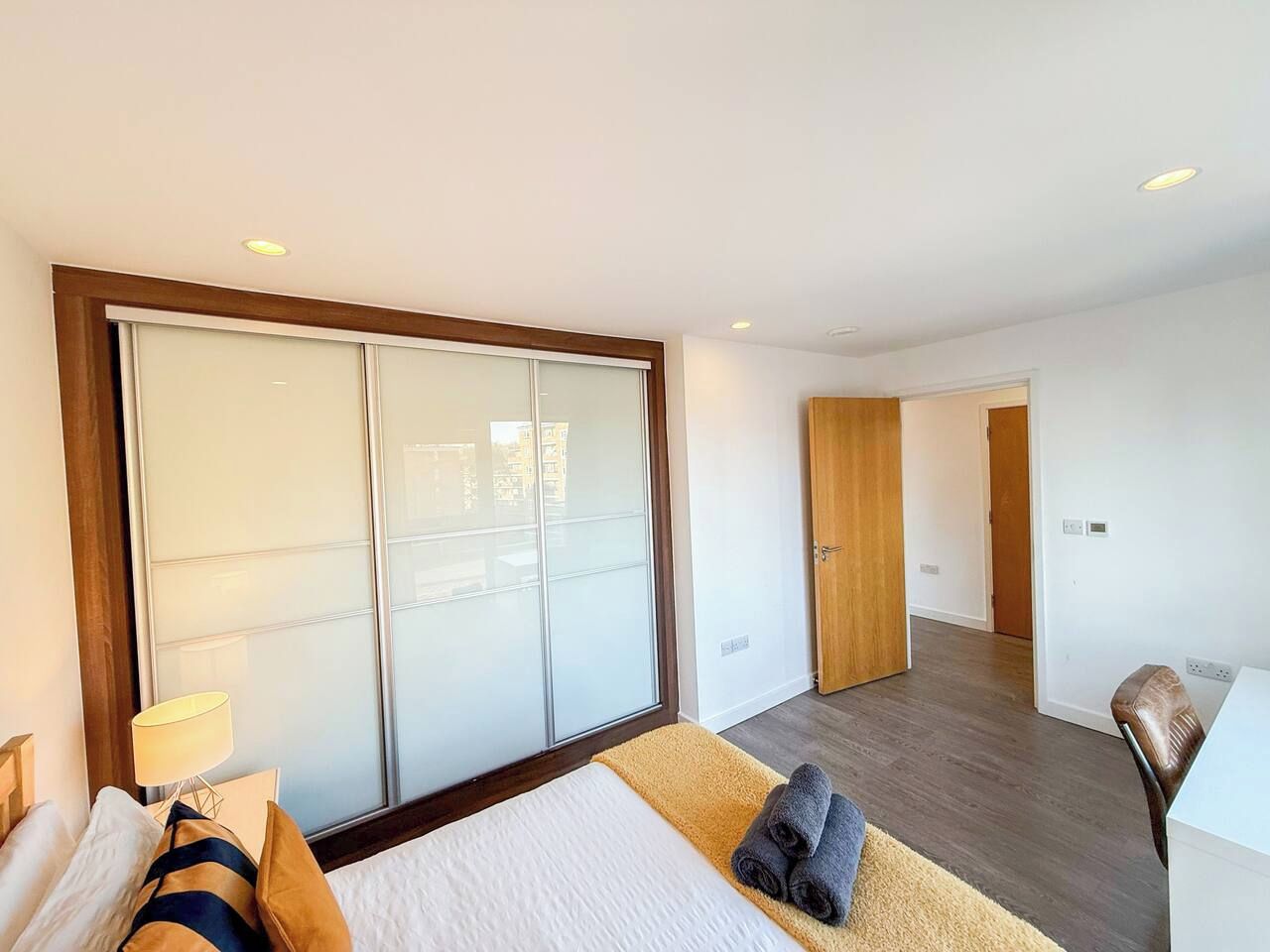 A modern hotel room with a large sliding door, a bed with a white and yellow bedspread, a small table with a lamp, and a chair.