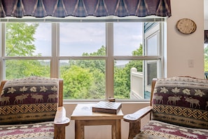 A room with two patterned armchairs facing a large window with a view of trees and a lake. A wooden table with a chessboard is in the center.
