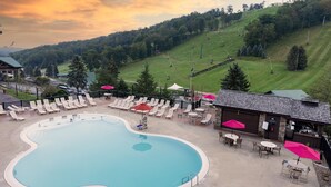 A swimming pool with lounge chairs and umbrellas, surrounded by a building with outdoor seating and a view of a hilly landscape.