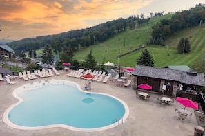 A swimming pool with lounge chairs and umbrellas, surrounded by a building with outdoor seating and a view of a hilly landscape.