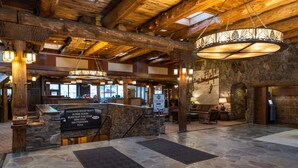 A spacious lobby with stone flooring, wooden beams, and a central chandelier. A reception desk is visible with a sign listing amenities like an alpine room and mini golf.