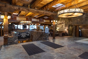 A spacious lobby with stone flooring, wooden beams, and a central chandelier. A reception desk is visible with a sign listing amenities like an alpine room and mini golf.