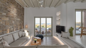 A modern living room with a stone wall, a sofa, a coffee table, and a TV. - Villa TROPICAL OASIS, Kalafatis, Mykonos. (Mykonos)