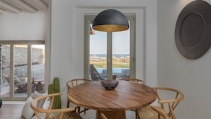 A dining area with a round wooden table, six chairs, a black pendant light, and a view through a glass door to the outdoors. - Villa TROPICAL OASIS, Kalafatis, Mykonos. (Mykonos)