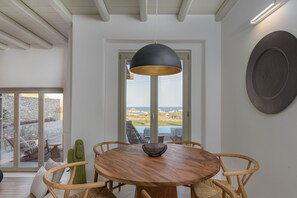 A dining area with a round wooden table, six chairs, a black pendant light, and a view through a glass door to the outdoors.
