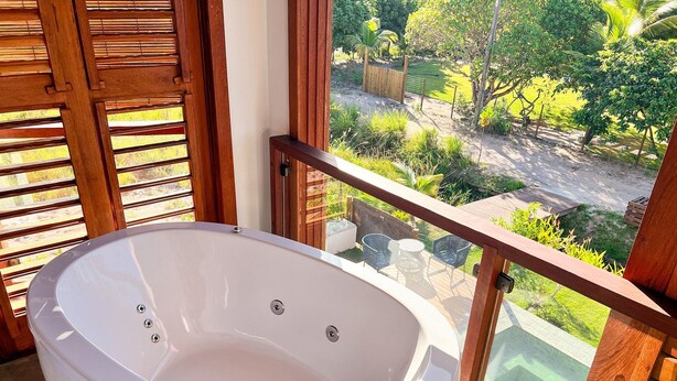 Una bañera blanca en un balcón con baranda de madera, con vistas a un jardín con árboles y césped.