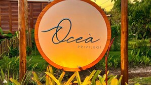 Un letrero con el nombre 'Ocea Privilegios' frente a una casa con contraventanas de madera y un jardín con plantas amarillas.