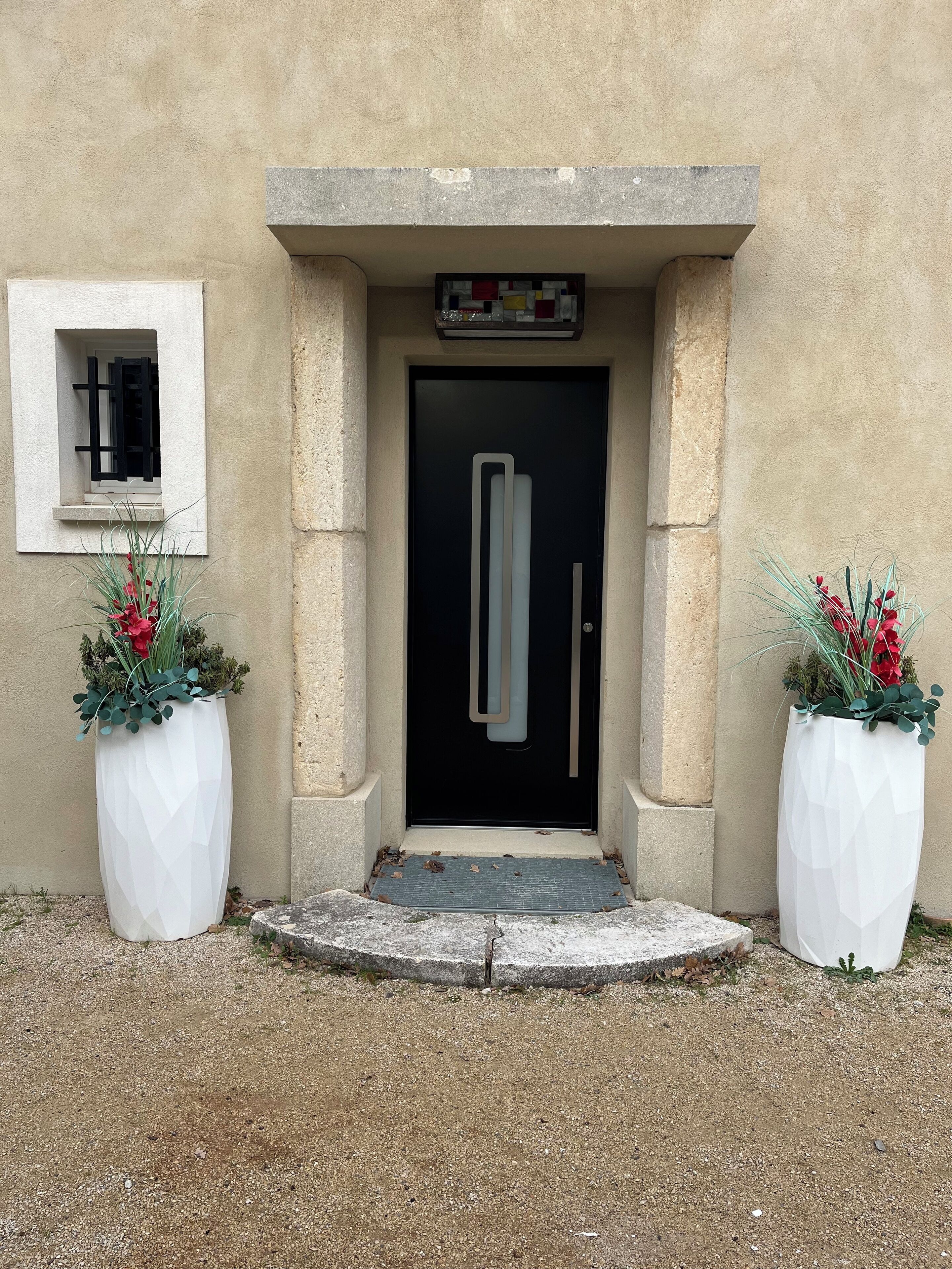 Ingresso di un edificio con una porta nera, una piccola finestra con persiane nere e due grandi fioriere bianche con fiori rossi.