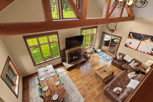 A spacious living area with a fireplace, a dining table set for four, a leather sofa, and large windows offering a view of the outdoors.