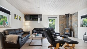 Een moderne woonkamer met een zwarte leren bank, een fauteuil en een salontafel. Er is een open haard met een stenen muur, een plafond met houten balken en een raam met zonwering.
