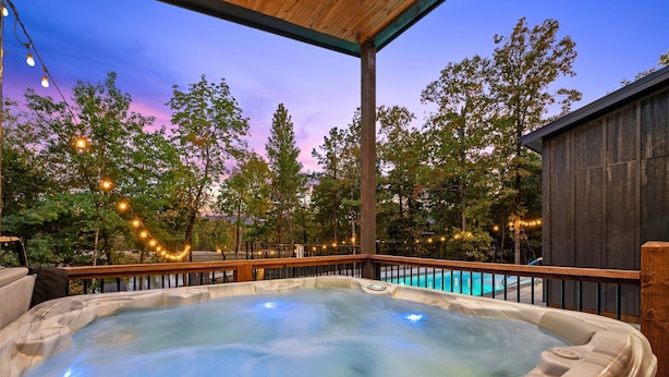 Uma banheira de hidromassagem com deck de madeira, cercada por árvores e luzes de corda, com uma piscina ao fundo.