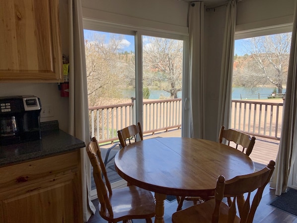 A wooden dining table with chairs in a room with large windows, a coffee maker, and a view of a lake and trees.