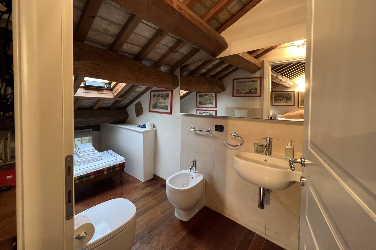 A compact bathroom with a bidet, toilet, and sink. It features wooden beams on the ceiling and a small window.