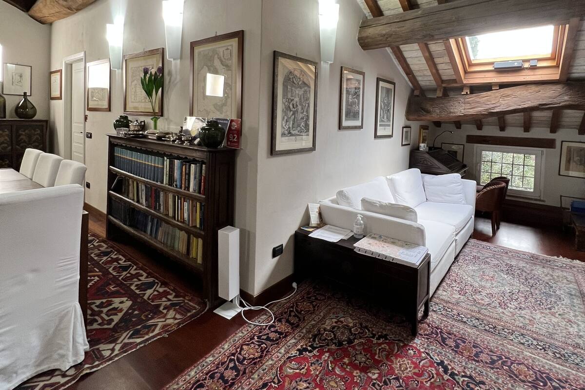 A spacious loft with high ceilings, exposed wooden beams, a skylight, a large bookshelf, a white sofa, and a patterned rug.
