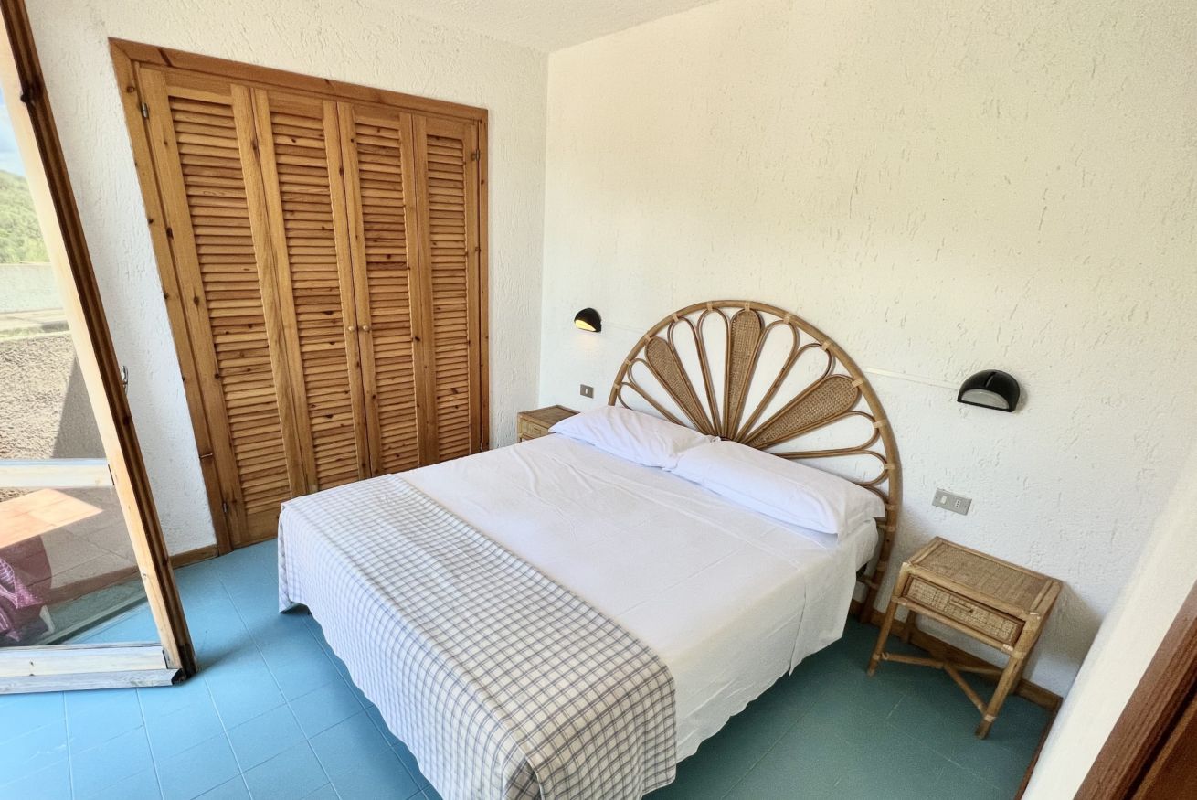 A bedroom with a large bed, a wooden wardrobe, a small bedside table, and a wall-mounted light.