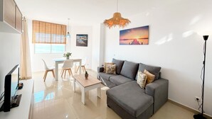 Una sala de estar moderna con un sofá gris, una mesa de comedor con sillas, un televisor y un amplio ventanal con persianas.