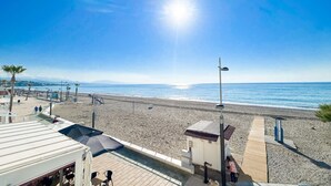 Vista frente al mar con cielo despejado, playa arenosa y un camino de madera que lleva a una pequeña construcción.