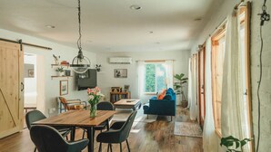 A spacious living area with a wooden dining table, black chairs, a blue sofa, and a large window with curtains. - Modern cottage centrally located with private access to the Rogue River! (Grants Pass)