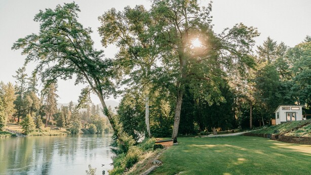 A serene lakeside property with a well-maintained lawn, a wooden dock, and a small house with a stone wall. - Modern cottage centrally located with private access to the Rogue River! (Grants Pass)