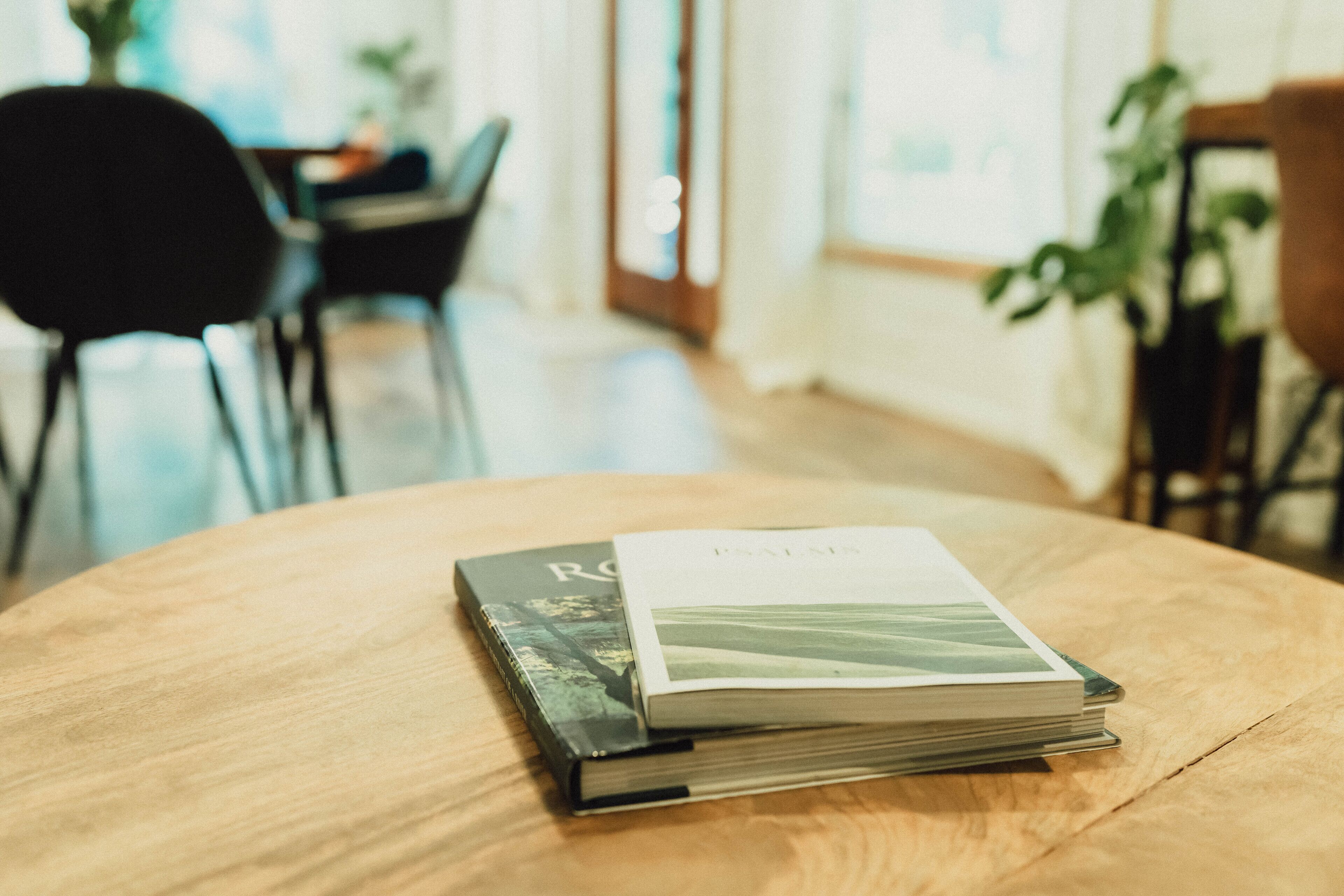Ein Stapel Bücher auf einem Holztisch in einem Raum, im Hintergrund ein Stuhl und eine Pflanze.