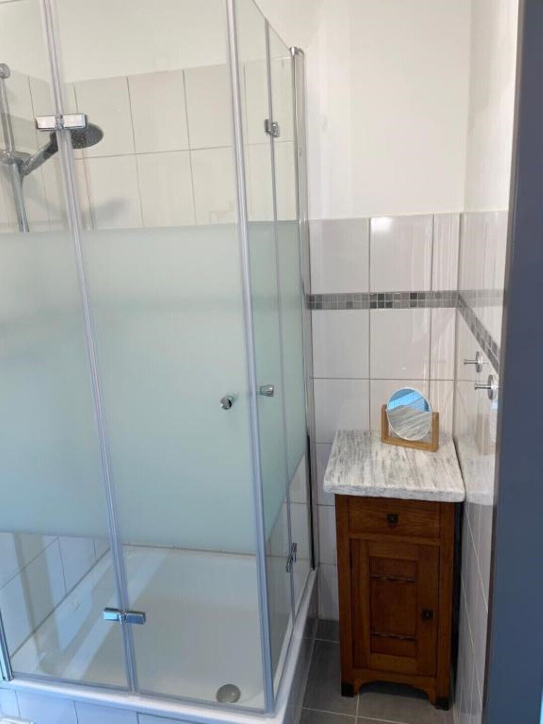 A bathroom with a glass shower enclosure, a wooden vanity with a mirror, and tiled walls.