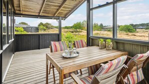 Una terraza de madera con una mesa y sillas, ventanas amplias y vistas del paisaje circundante.