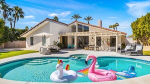 A swimming pool with inflatable unicorn and flamingo floats, surrounded by a house with a patio and outdoor furniture. - Presidential Vacation Retreat (Rancho Mirage)
