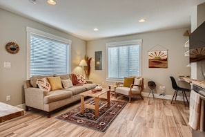 A living room with a sofa, coffee table, and chairs. There are two windows with blinds, a lamp, and a wall-mounted artwork.