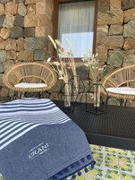 A stone wall exterior with two wicker chairs and a small table, a striped blue and white cushion, and a vase with dried plants.
