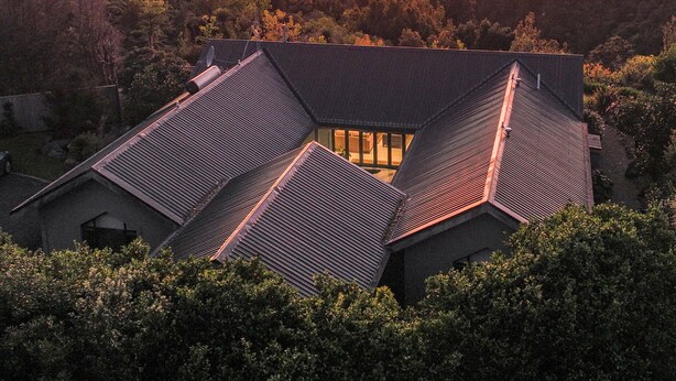 Une maison moderne, dotée d’un toit en pente, entourée d’une forêt dense et offrant une vue sur un plan d’eau au coucher du soleil.