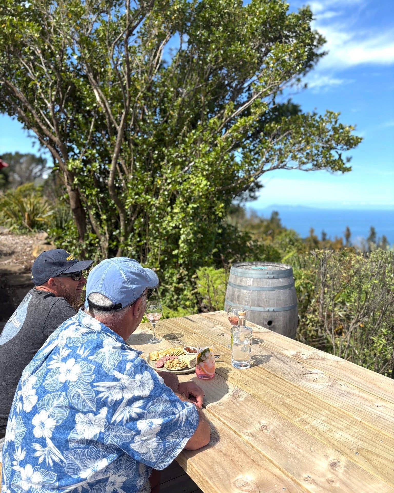 Une table en bois, dressée à l’extérieur, avec un repas et des boissons, des arbres en arrière-plan et un ciel dégagé.