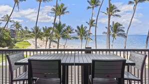Een houten terras met een tafel en stoelen met uitzicht op een strand met palmbomen en de oceaan.